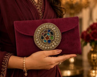 May include: A deep burgundy velvet clutch bag featuring a large, circular central embellishment. The ornament is crafted with a gold-toned border, radiating clear crystals, and a cluster of multi-colored, polished stones in the center.