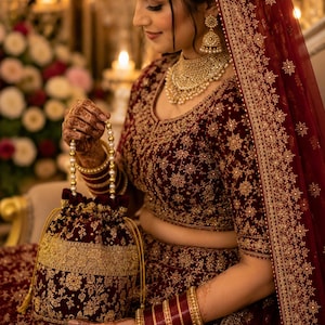 May include: A deep maroon velvet drawstring handbag featuring intricate gold floral embroidery and a beaded pearl handle. The bag is adorned with a wide gold metallic band and matching gold drawstring cords, showcasing a rich, textured finish.