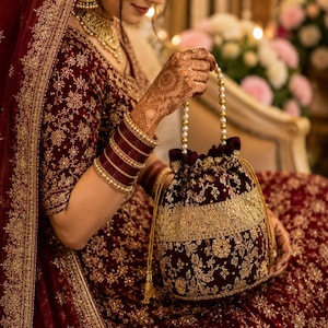 May include: A deep maroon velvet drawstring pouch featuring intricate gold floral embroidery and a wide metallic gold band. The bag includes a pearl-beaded handle and gold cord drawstrings with tassels, matching the ornate aesthetic of the attire.