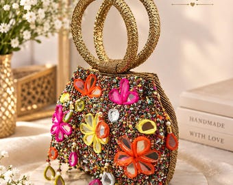 May include: A structured handbag with two circular gold handles, featuring a dark base densely covered in multicolored beads and reflective mirror accents shaped like flowers. Dangling beaded fringe with small mirror charms hangs from the bottom.