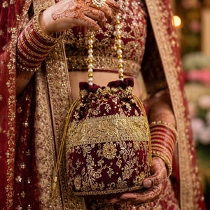 May include: A deep maroon velvet potli bag featuring intricate gold embroidery and a drawstring closure. The bag is accented with a handle made of white pearl beads and gold-toned spacers, matching the ornate floral patterns on the fabric.