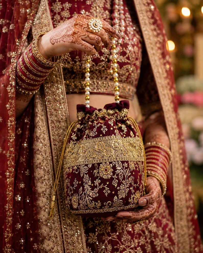 May include: A deep maroon velvet potli bag featuring intricate gold embroidery and a drawstring closure. The bag is accented with a handle made of white pearl beads and gold-toned spacers, matching the ornate floral patterns on the fabric.