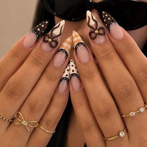 Pode incluir: Unhas pontiagudas em formato de amêndoa com base de manicure francesa, pontas em estilo renda preta, bolinhas marrons e brancas e padrões xadrez. Dois laços marrons tridimensionais com centros dourados estão aplicados. Anéis dourados são usados.