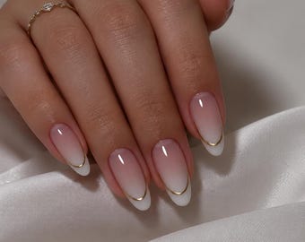 May include: Almond-shaped nails featuring a soft pink to white ombre gradient finish. Each nail is accented with a thin, curved gold line detail along the white tip. A delicate gold ring with a small clear stone is worn on the ring finger.