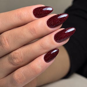 May include: Almond-shaped fingernails featuring a deep, dark red polish with a fine, shimmering glitter finish. The nails have a smooth, glossy top coat that reflects light, creating a polished and uniform appearance across all four visible nails.