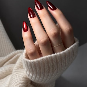 May include: A hand with long, almond-shaped nails painted in a glossy, deep metallic red finish. The fingers are adorned with a thin, plain gold-toned band ring on the ring finger. The hand is partially covered by a cream-colored ribbed knit sweater.