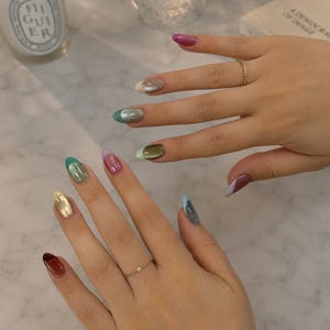 以下が含まれることがあります： アーモンド型の爪にマルチカラーのフレンチマニキュアを施した両手。各爪の先端はピンク、シルバー、グリーン、ゴールド、ブラウン、ブルーなどのメタリックや光沢のある色で彩られ、2つの細いゴールドリングがアクセントになっています。