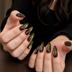 May include: Almond-shaped fingernails featuring a cat-eye polish effect with a deep, dark base color transitioning into a shimmering, metallic olive green center. The finish is glossy and reflective, creating a distinct light-catching streak across each nail.