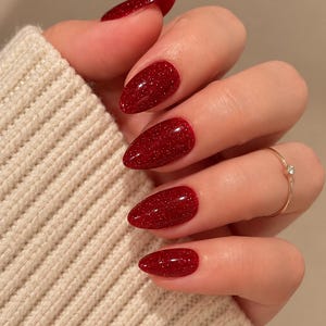 May include: Almond-shaped fingernails painted with a glossy, deep red polish containing fine silver glitter particles. A thin gold ring with a small clear stone is worn on the ring finger, and a cream-colored ribbed knit sleeve is visible.