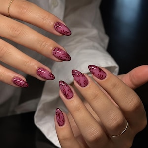 May include: Almond-shaped nails featuring a translucent mauve base color with dark burgundy floral patterns painted on each nail. The design includes delicate leaf and petal motifs that follow the curve of the nail bed, finished with a glossy top coat.