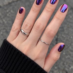 May include: A silver wavy ring and a thin silver band worn on fingers with short, rounded nails featuring a reflective, metallic chrome finish that shifts between deep purple, blue, and magenta tones against a black ribbed sleeve.