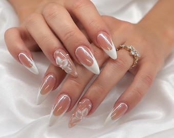 May include: Almond-shaped press-on nails featuring a classic French manicure design with white tips and a nude base. Two nails are accented with three-dimensional white floral petals and small white pearls. A gold-toned ring with clear stones.