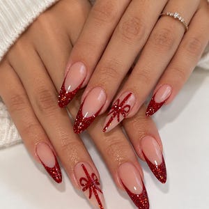 Könnte beinhalten: Lange, mandelförmige Nägel mit einer nudefarbenen Basis und glitzernden roten French-Tips. Zwei der Nägel sind mit einem handgemalten roten Glitzerschleifen-Design akzentuiert, das an ein Geschenkband erinnert und eine detaillierte Maniküre ergibt.