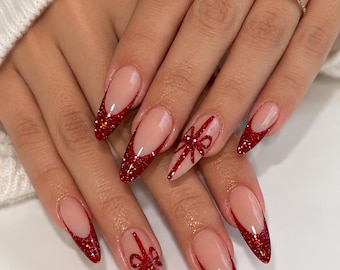 May include: Long almond-shaped nails featuring a nude base with sparkling red glitter French tips. Two of the nails are accented with a hand-painted red glitter bow design that resembles a gift wrap ribbon, creating a festive and detailed manicure.