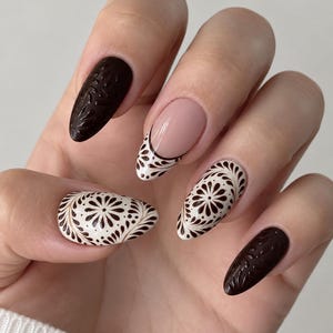 May include: Almond-shaped nails featuring a mix of solid dark brown with embossed patterns, cream-colored nails with intricate dark brown floral designs, and a French manicure style with a cream base and dark brown floral detailing at the tip.