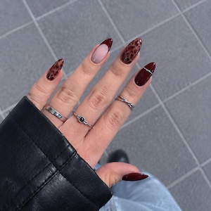 Könnte beinhalten: Mandelformige Fingernägel mit einer Kombination aus tiefrotem Burgunder-Nagellack, Leopardenmuster und French-Maniküre-Design. Die Finger sind mit drei silberfarbenen Ringen geschmückt, darunter ein schlichter Ring, ein Steinring und ein dünner Kreuzring.