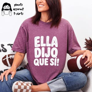 May include: A maroon t-shirt with the text "ELLA DIJO QUE SÍ!" in white. The shirt is worn with blue jeans. The image also includes a football and the text "planets different t-shirts".
