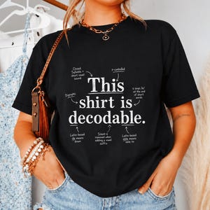 Op de afbeelding: Een zwart t-shirt met korte mouwen en de witte tekst This shirt is decodable. Het ontwerp bevat taalkundige annotaties met pijlen die naar specifieke woorden wijzen, waarbij concepten zoals gesloten lettergrepen en Latijnse wortels worden uitgelegd.