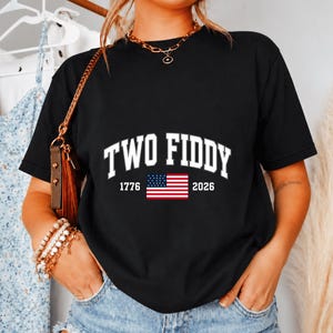 Könnte beinhalten: Ein schwarzes kurzärmeliges T-Shirt mit dem weißen Schriftzug TWO FIDDY in einer College-Schriftart, wobei die Zahlen 1776 und 2026 eine kleine Grafik der Flagge der Vereinigten Staaten in Rot, Weiß und Blau auf der Brustmitte flankieren.