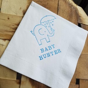 May include: White square paper napkin with a blue illustration of an elephant holding an umbrella with heart shapes. The text "BABY HUNTER" is printed below the elephant.