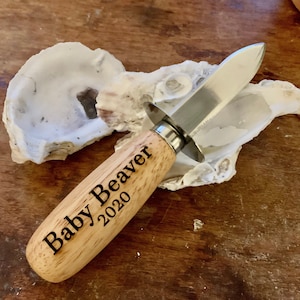 May include: An oyster knife with a wooden handle engraved with "Baby Beaver 2020" and a stainless steel blade. The knife rests on an oyster shell, with a wooden surface in the background.