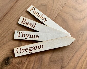 May include: Four wooden garden markers with the names "Parsley", "Basil", "Thyme", and "Oregano" etched in a dark brown font. The markers are arranged on a wooden surface.