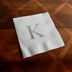May include: A white paper napkin with a gray letter "K" printed on it. The napkin is placed on a dark brown wooden surface with a geometric pattern. The napkin has a textured border.
