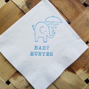 May include: White square napkin with a blue elephant illustration holding an umbrella with heart-shaped raindrops. The text "BABY HUNTER" is printed below the elephant. The napkin has a ribbed border.