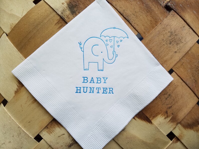 May include: White square napkin with a blue elephant illustration holding an umbrella with heart-shaped raindrops. The text "BABY HUNTER" is printed below the elephant. The napkin has a ribbed border.