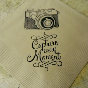 May include: A beige napkin with a black and white illustration of a vintage camera. The text "Capture every Moment" is written in elegant script below the camera. The napkin has a ribbed border.