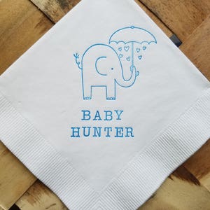 May include: White square napkin with a blue elephant holding an umbrella with heart-shaped raindrops. The text "BABY HUNTER" is printed in blue below the elephant. The napkin has a ribbed border.