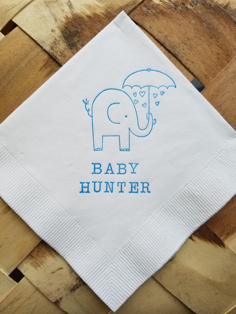 May include: White square napkin with a blue elephant holding an umbrella with heart-shaped raindrops. The text "BABY HUNTER" is printed in blue below the elephant. The napkin has a ribbed border.