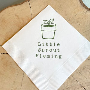 May include: White paper napkin featuring a green illustration of a seedling in a pot. The text "Little Sprout Fleming" is printed beneath the illustration in green. The napkin is displayed on a wooden surface.