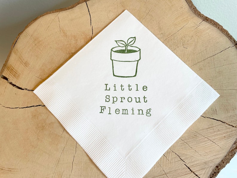 May include: White paper napkin featuring a green illustration of a seedling in a pot. The text "Little Sprout Fleming" is printed beneath the illustration in green. The napkin is displayed on a wooden surface.