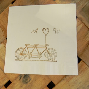 May include: A cream-colored paper napkin with a tandem bicycle illustration in a gold tone. The design includes the letters "A" and "W" with a heart-shaped balloon. The napkin is square and has a textured surface.