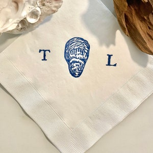 May include: White square linen napkin with a blue oyster print in the center, flanked by the letters "T" and "L". The napkin has a white ribbed border. An oyster shell and driftwood are in the background.