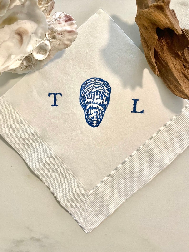 May include: White square linen napkin with a blue oyster print in the center, flanked by the letters "T" and "L". The napkin has a white ribbed border. An oyster shell and driftwood are in the background.