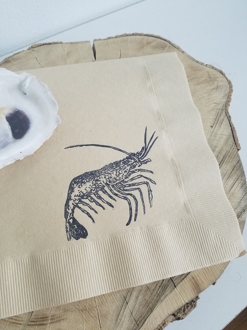Pode incluir: Guardanapo de papel bege com uma ilustra&ccedil;&atilde;o de lagosta preta. O guardanapo tem uma borda canelada e &eacute; colocado em uma fatia de madeira. Uma concha de ostra branca est&aacute; no canto superior esquerdo.