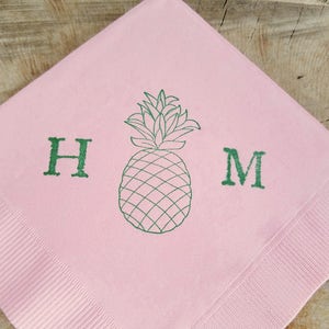 May include: Pink paper napkin with a green pineapple illustration and the letters "H" and "M". The napkin has a ribbed texture on the edges and is set against a wooden background.