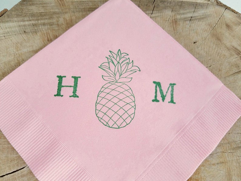 May include: Pink paper napkin with a green pineapple illustration and the letters "H" and "M". The napkin has a ribbed texture on the edges and is set against a wooden background.