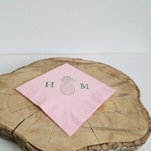 May include: A pink paper napkin with a green pineapple illustration and the letters "H" and "M". The napkin is placed on a wooden surface with a white background.