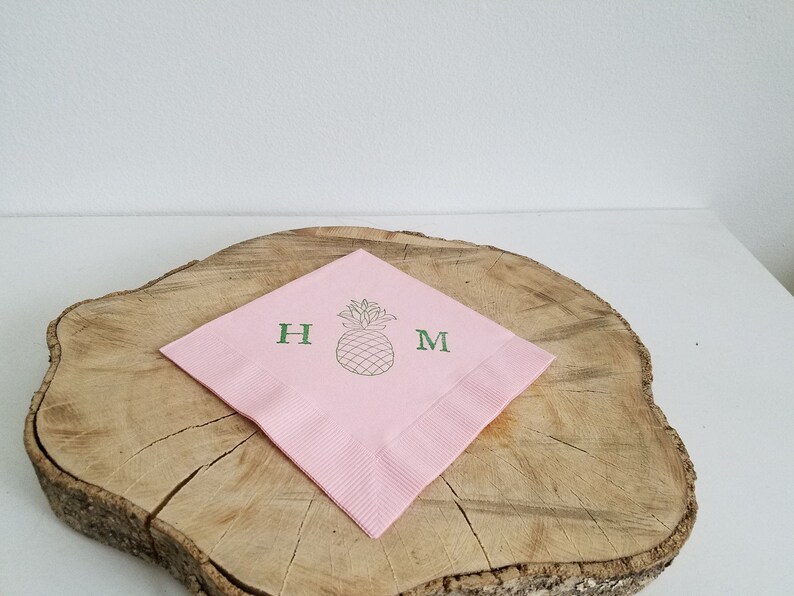 May include: A pink paper napkin with a green pineapple illustration and the letters "H" and "M". The napkin is placed on a wooden surface with a white background.