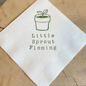 May include: White square napkin with a green illustration of a potted plant and the words "Little Sprout Fleming" printed in green. The napkin has a ribbed border and is displayed on a wooden surface.