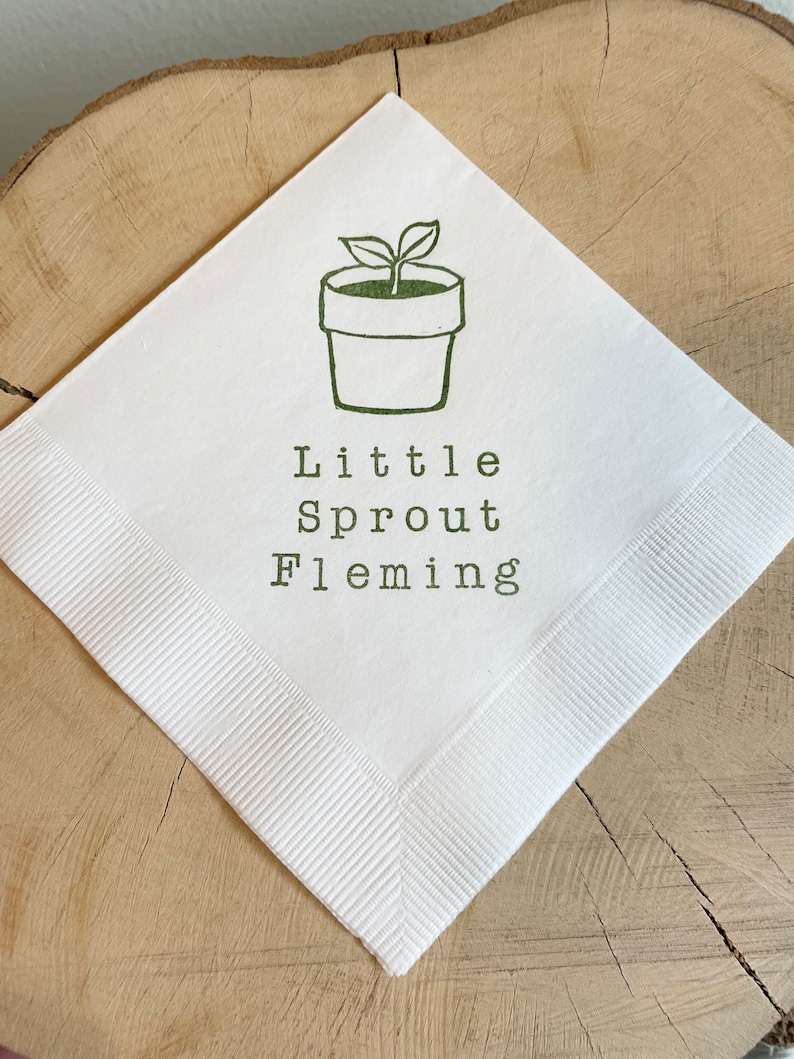 May include: White square napkin with a green illustration of a potted plant and the words "Little Sprout Fleming" printed in green. The napkin has a ribbed border and is displayed on a wooden surface.
