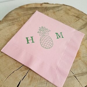 May include: A square, light pink napkin with a green pineapple illustration and the letters "H" and "M". The napkin has a ribbed border and is displayed on a wooden surface.