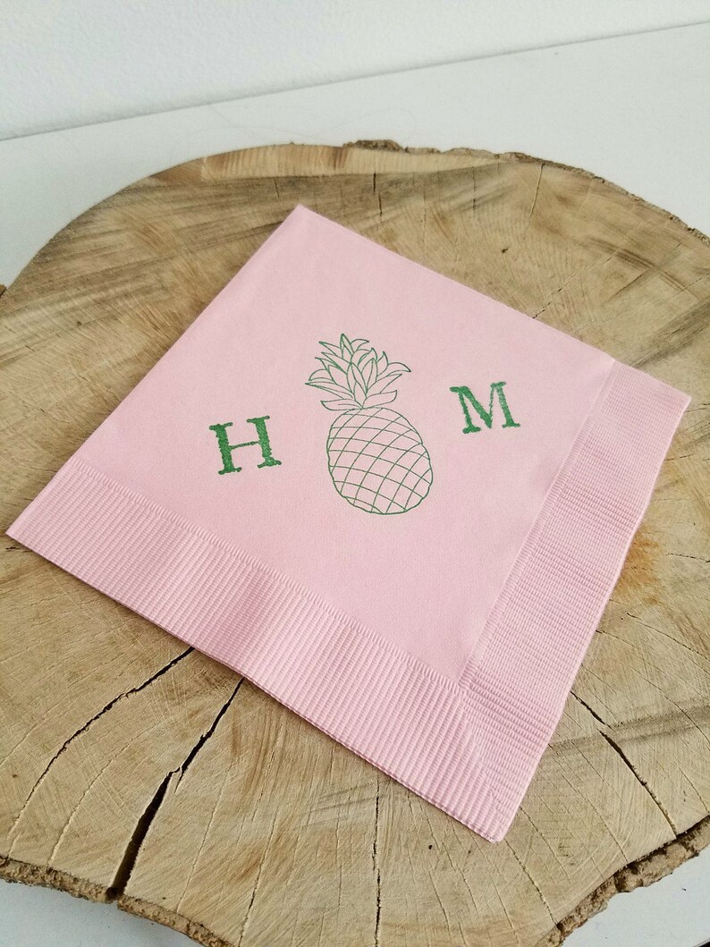 May include: A square, light pink napkin with a green pineapple illustration and the letters "H" and "M". The napkin has a ribbed border and is displayed on a wooden surface.