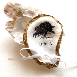 May include: A decorative oyster shell ring dish with a gold-painted edge. The white interior features a black tree illustration and the initials "D & A". A white ribbon is tied around the shell.