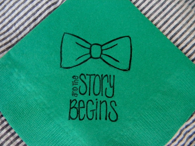 Op de afbeelding: Een vierkante, smaragdgroene servet met een zwarte vlinderdasillustratie en de woorden "and The STORY BEGINS" in een handgeschreven lettertype. De servet ligt op een gestreepte stoffen achtergrond.