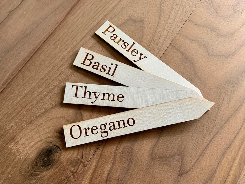 Op de afbeelding: Vier houten tuinmarkeringen met de namen "Peterselie", "Basilicum", "Tijm" en "Oregano" in donkerbruine letters. De markeringen zijn gerangschikt op een houten oppervlak.