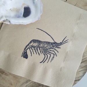 Pode incluir: Um guardanapo de papel bege com uma ilustra&ccedil;&atilde;o de camar&atilde;o preto, colocado em uma fatia de madeira. Uma concha de ostra branca vazia est&aacute; no canto superior esquerdo. O guardanapo tem uma borda canelada.
